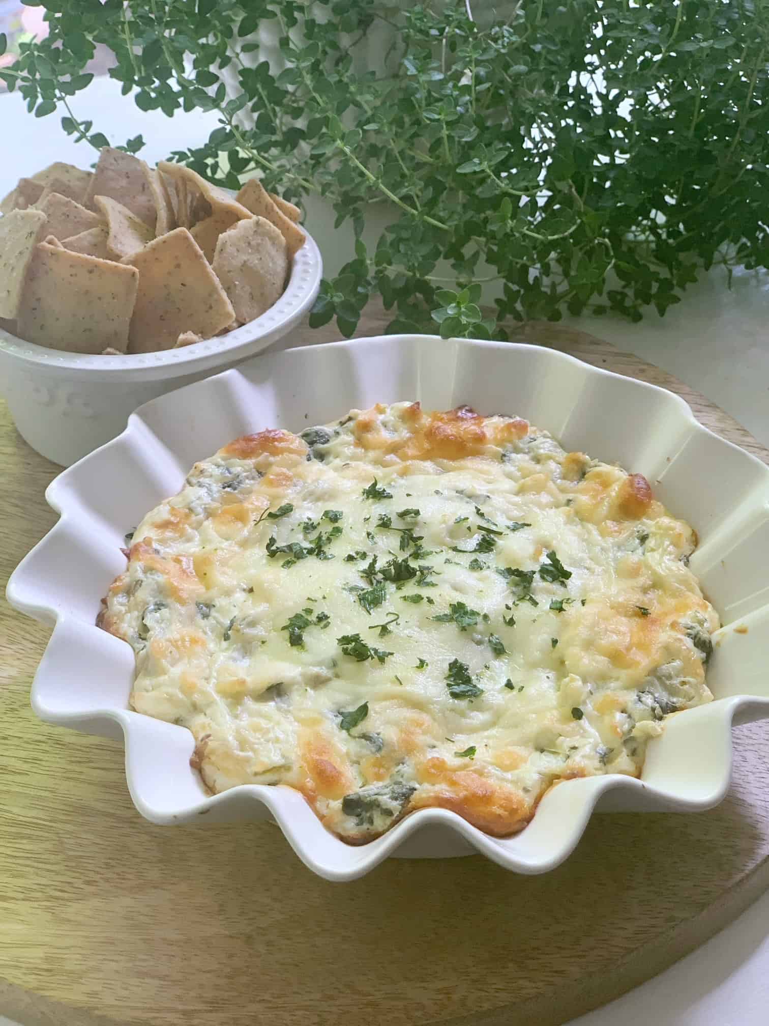 Creamy, cheesy goodness! A white ruffle edge baking dish with this no mayo spinach and artichoke dip. It is on a serving board with crackers. 