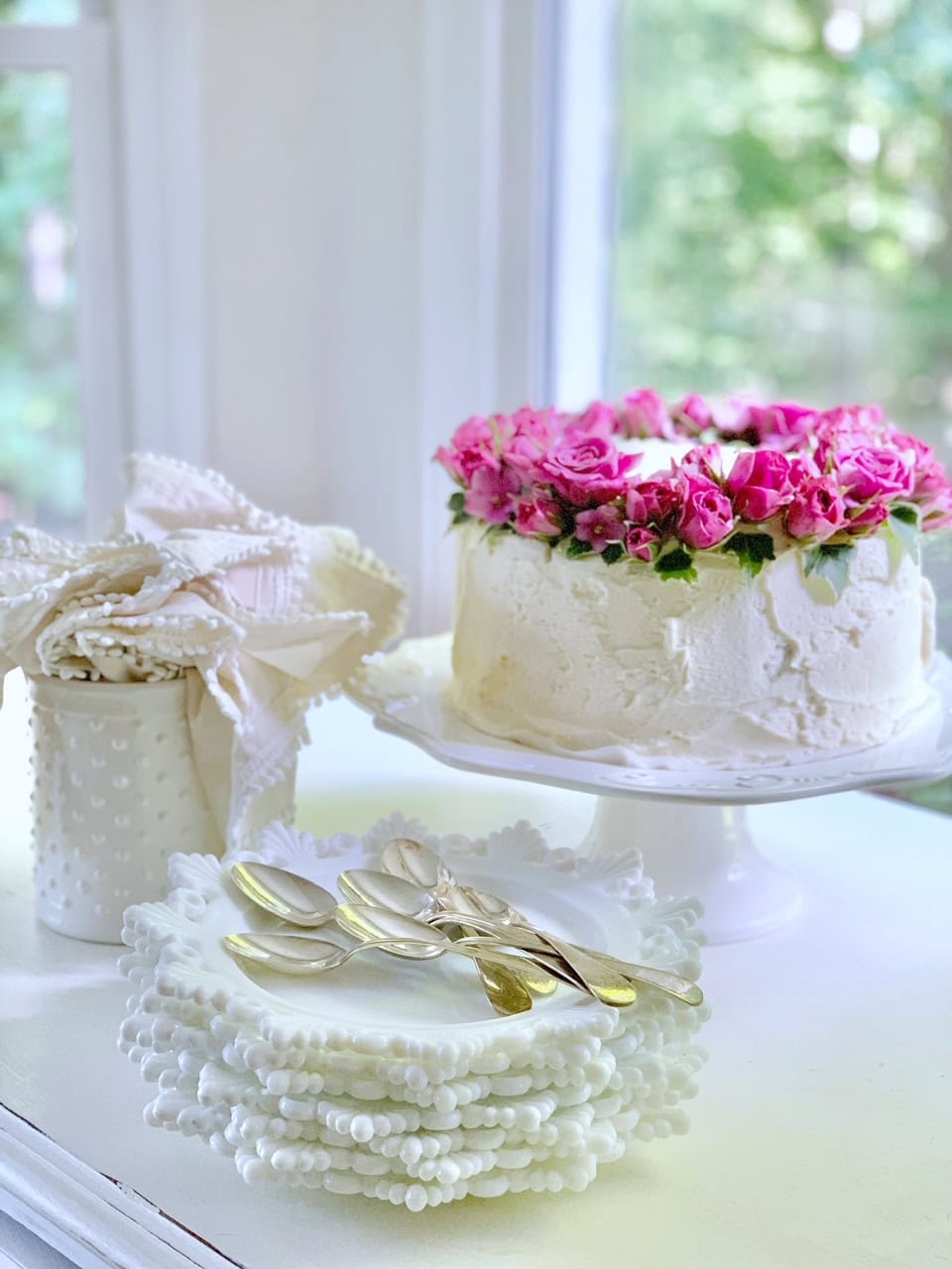 The cake with fresh flowers on a cake stand. There are cloth napkins, plates and silver spoons. 
