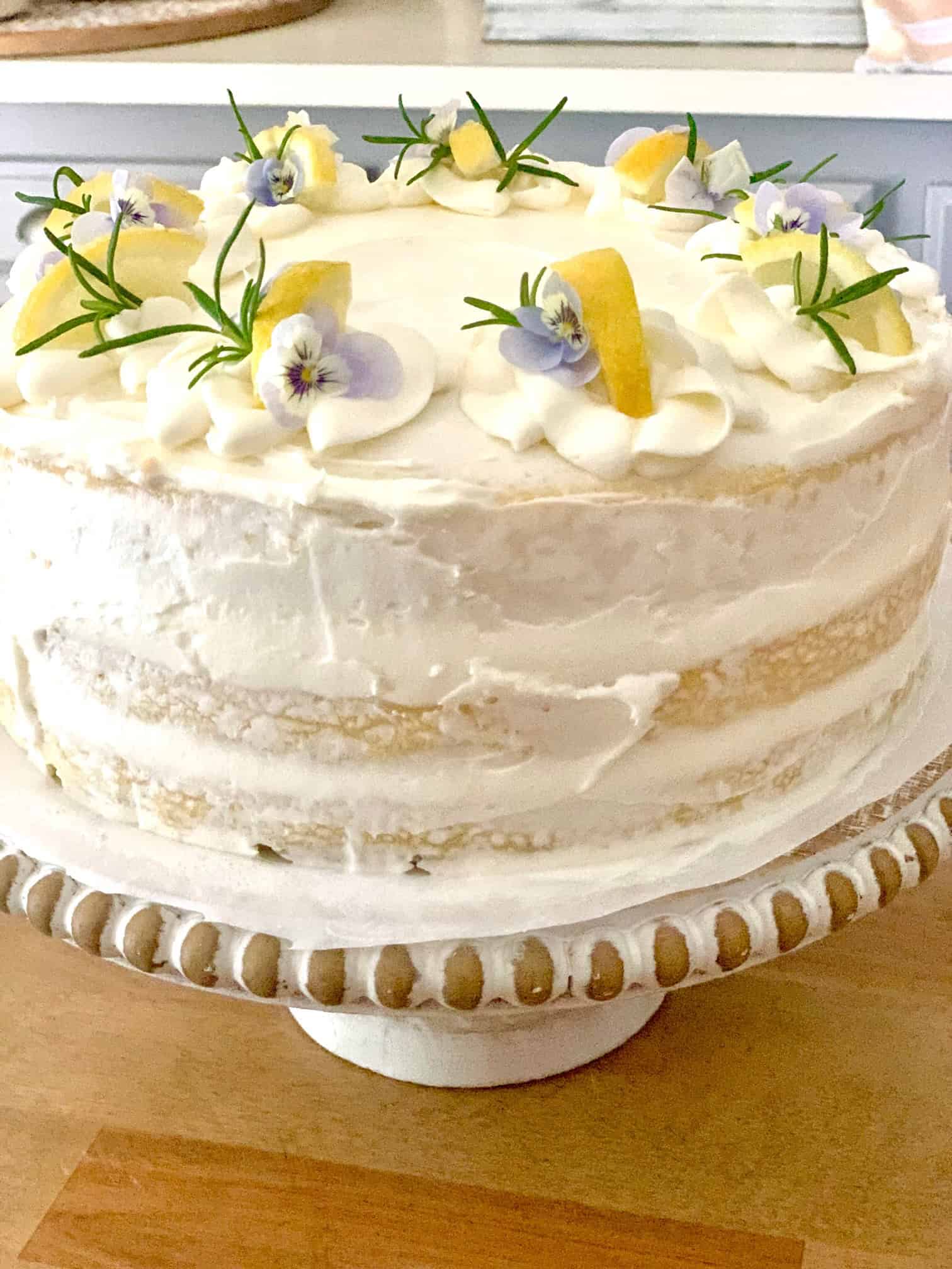 A lemon cake with white frosting. The top is decorated with lemon slices, rosemary and pansies. 