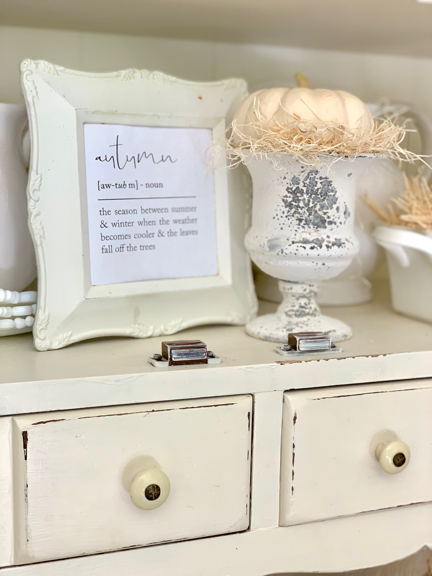 A shelf on a hutch with a frames fall saying and pumpkins in compotes.