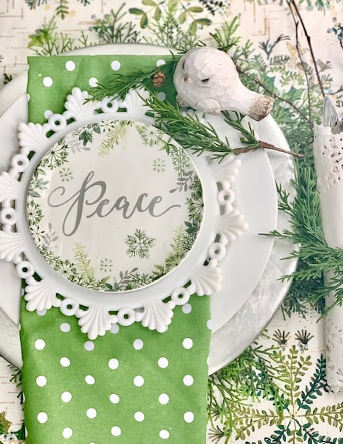 A green and white place setting on a silver charger. the top dish reads "peace" and there is a white wooden bird at the place setting. 