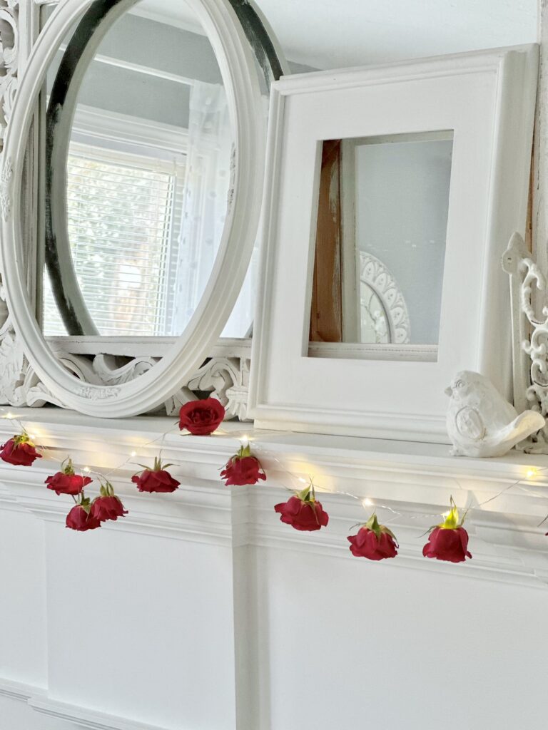 a straight on view of the rose garland on a mantel with a mirror, white frames and a bird. 