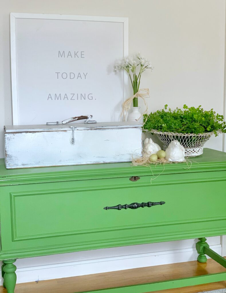 A chalk painted green blanket chest with an antique tool box and plants on top. 