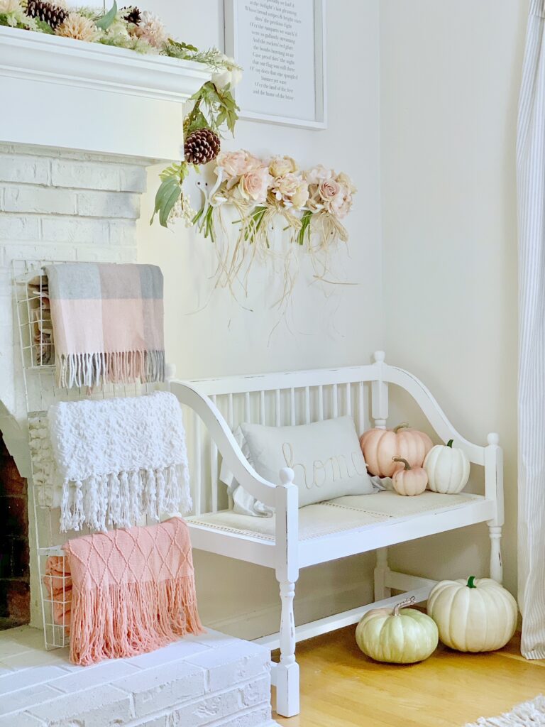 Coral, white and gray blankets spilling out of a white wire magazine rack in front of a fireplace. There is a white bench and white and coral pumpkins. 