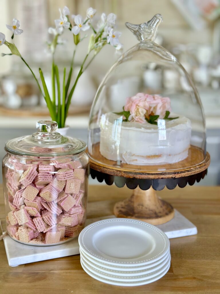 a cake stand with a cake that has hydrangea on top. canister of cookies and a stack of plates. 