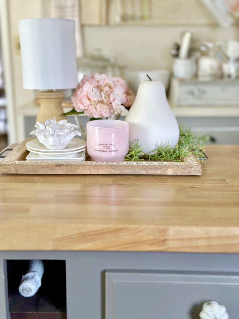 a wooden tray sitting on our island with a lamp, pink hydrangea, candles, a large white ceramic pear and some greens. 