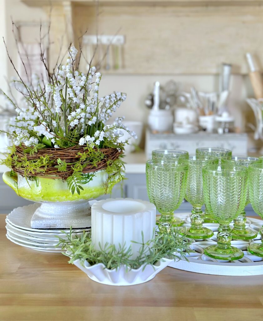a faux lily of the valley birds nest arrangement in a green and gray footed vase with green goblet style glasses and a candle. 