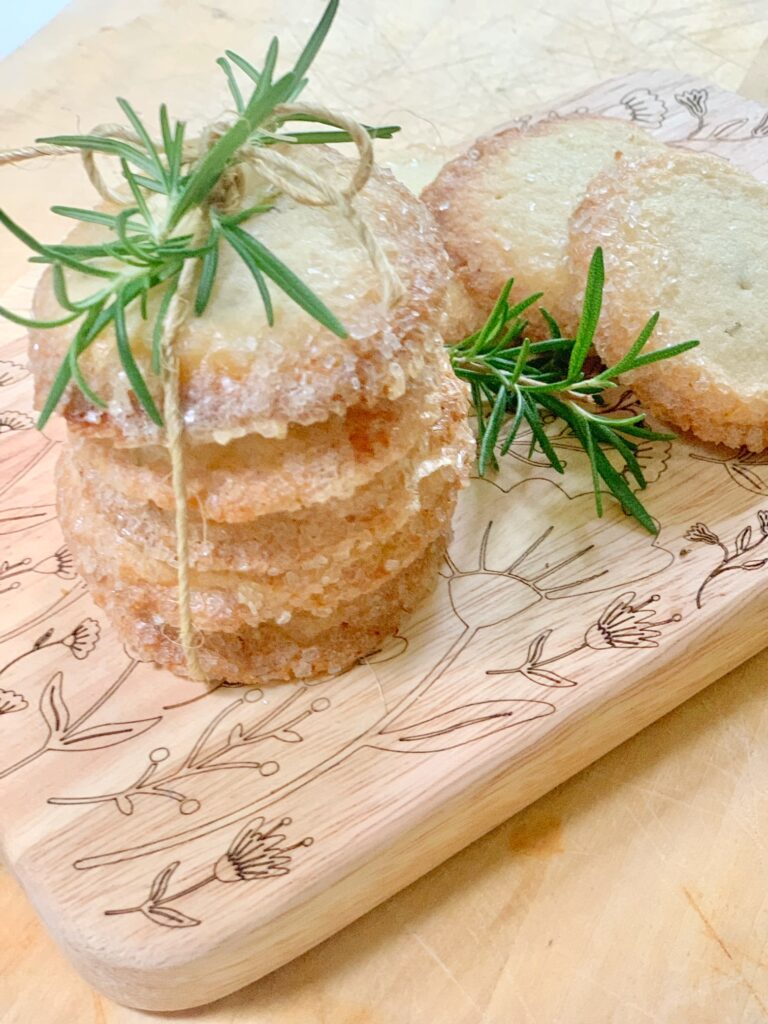 Rosemary butter cookies with a sprig of fresh rosemary on top. These cookies are stacked and on a cutting board with etched flowers. 