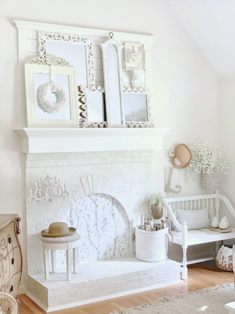 Full view of the fireplace. The fireplace is painted white with the layered mirrors sitting on the mantel. Also seen in the photo is a stool, basket of birch logs, a white antique bench and a hat. 