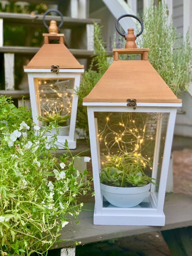 Two lanterns that are white with a warm honey stained top with a bowl of succulents inside on the bottom and twinkle lights floating to look like lightning bugs.