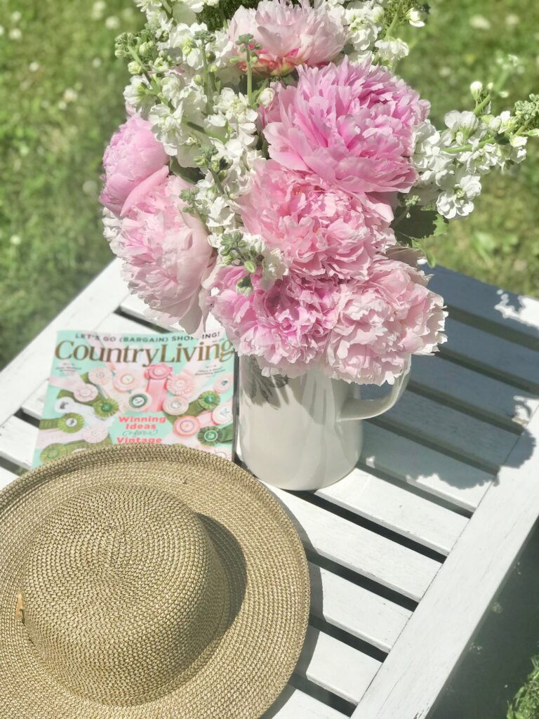 A pitcher filled with pink peonies sitting on an outdoor table with a hat and magazine. 