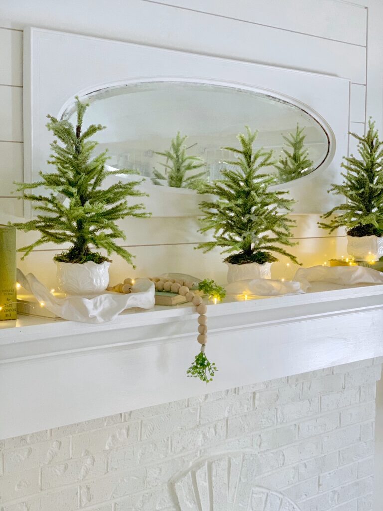 white mantel with mirroe with faux mini Christmas trees running across with decor beads. 