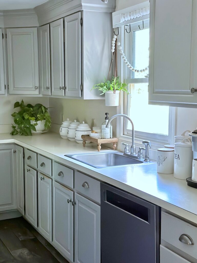 long view of the grey cabinets with off white counters, sink, dishwasher, window and decor. 