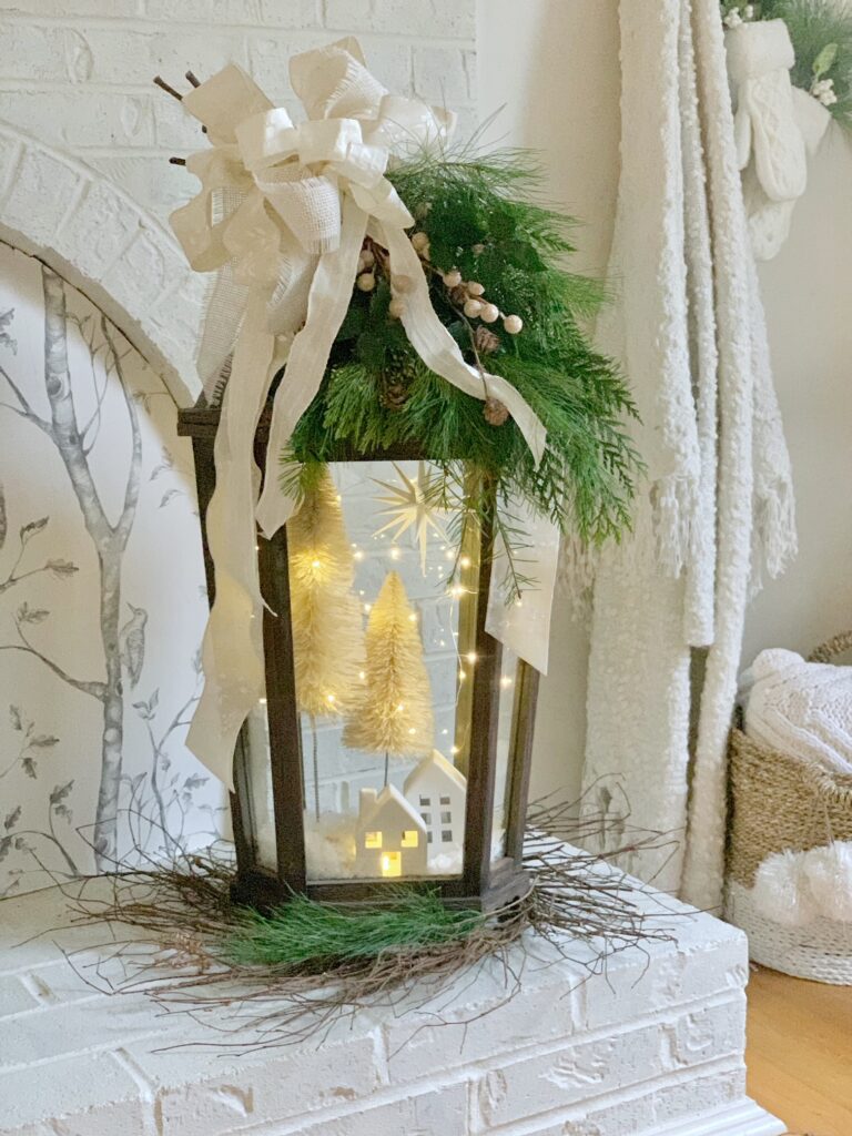 Lantern decorated  with bottle brush trees, twinkle lights with greenery swag and a large layered white bow. 