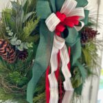 a pine christmas wreath with pinecones and a large bow that is made with a wide green ribbon, beige strip ribbon and a red small ribbon.