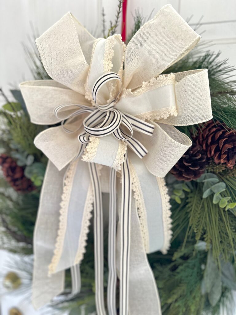 Large pine wreath with a large over the top cream colored bow that is made from 3 different ribbons. 