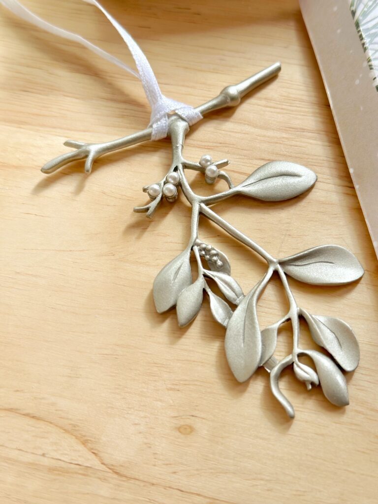 A photo of just the ornament. it is a brushes silver ornament shaped like a delicate sprig of mistletoe. It has a thin white ribbon for hanging. 