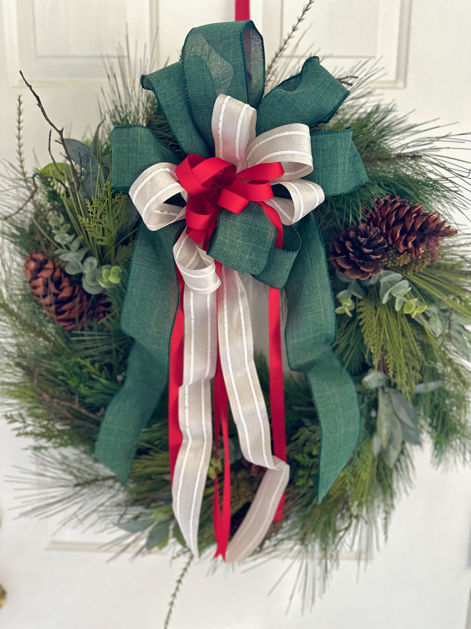 A front view of a pine wreath with a green, cream and red layered bow.