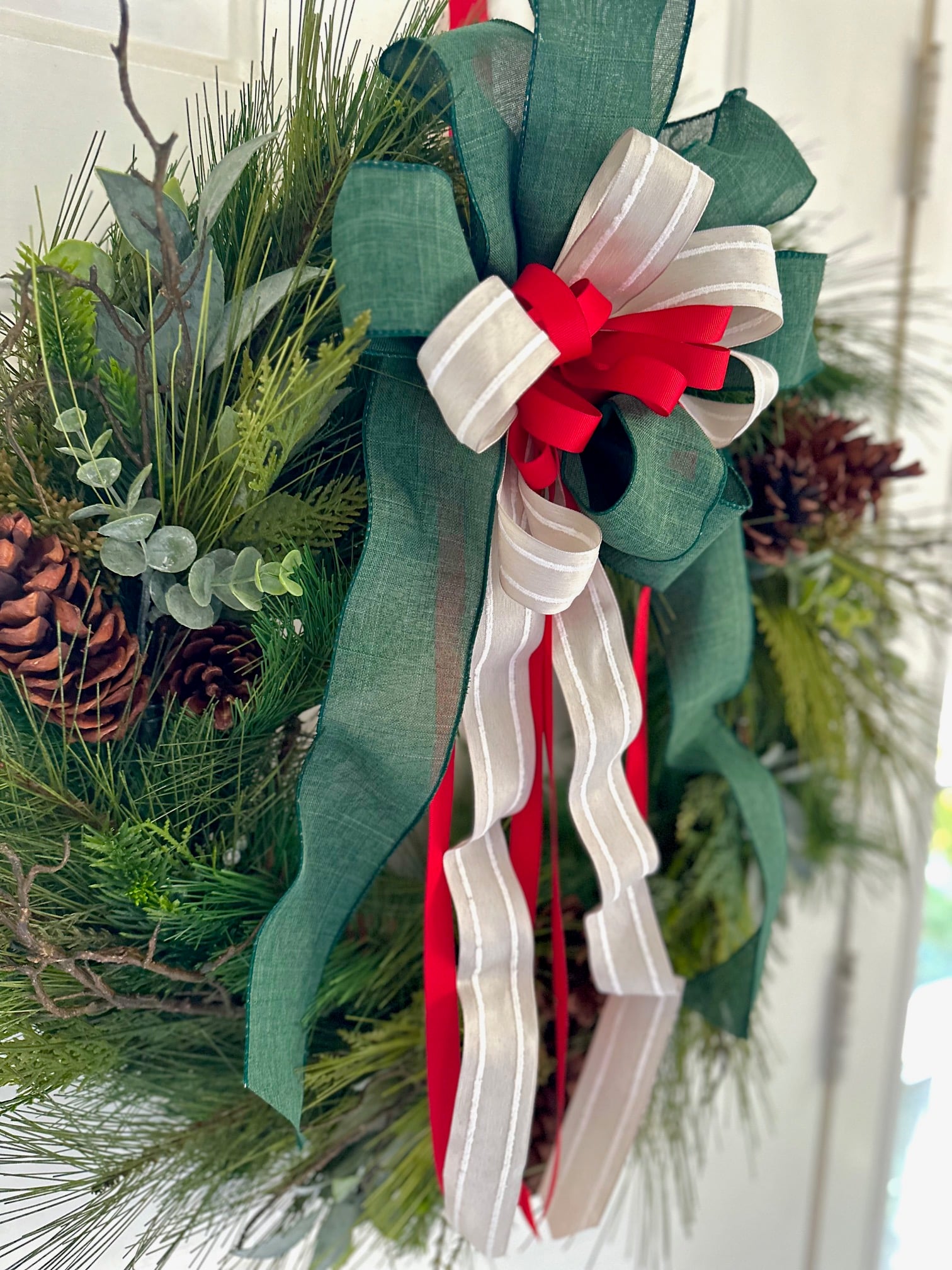 A pine Christmas wreath with pinecones and a large bow that is made with a wide green ribbon, beige strip ribbon and a red small ribbon.