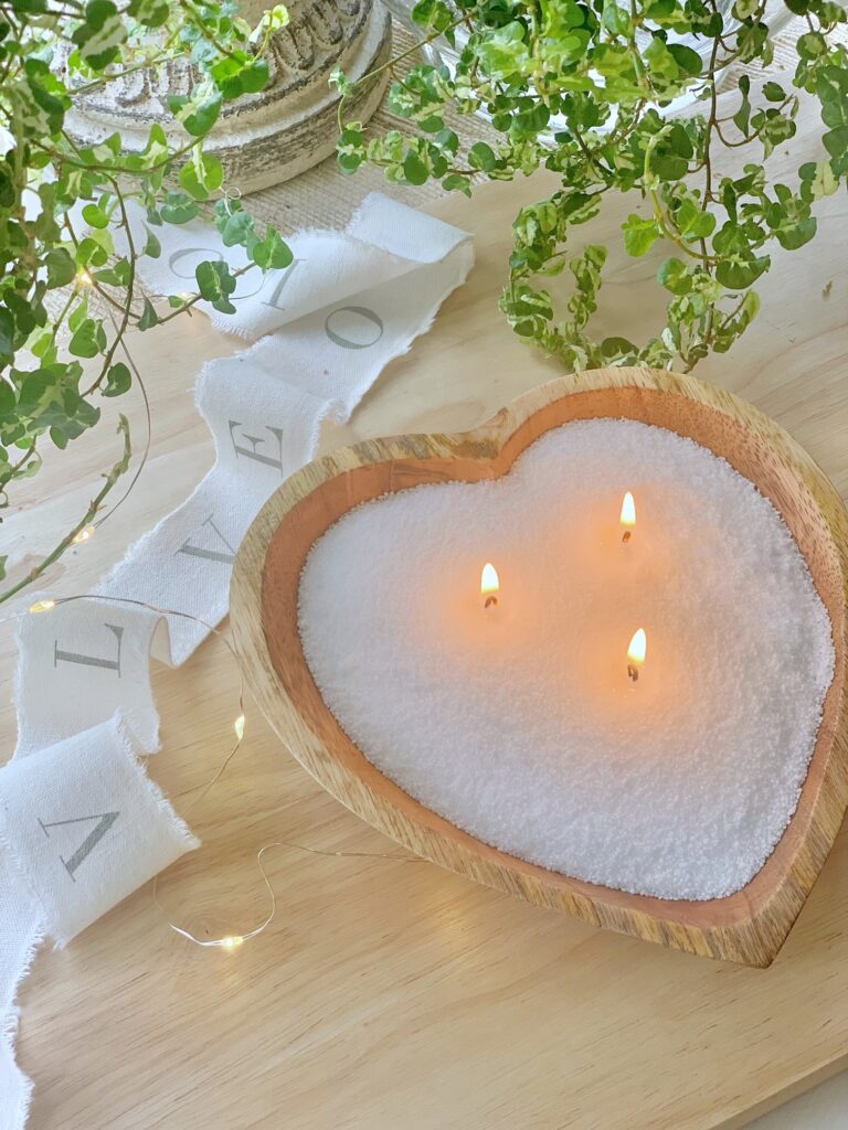 A wooden heart-shaped bowl filled with pearled candle on a wood board with a ribbon that reads "Love". 