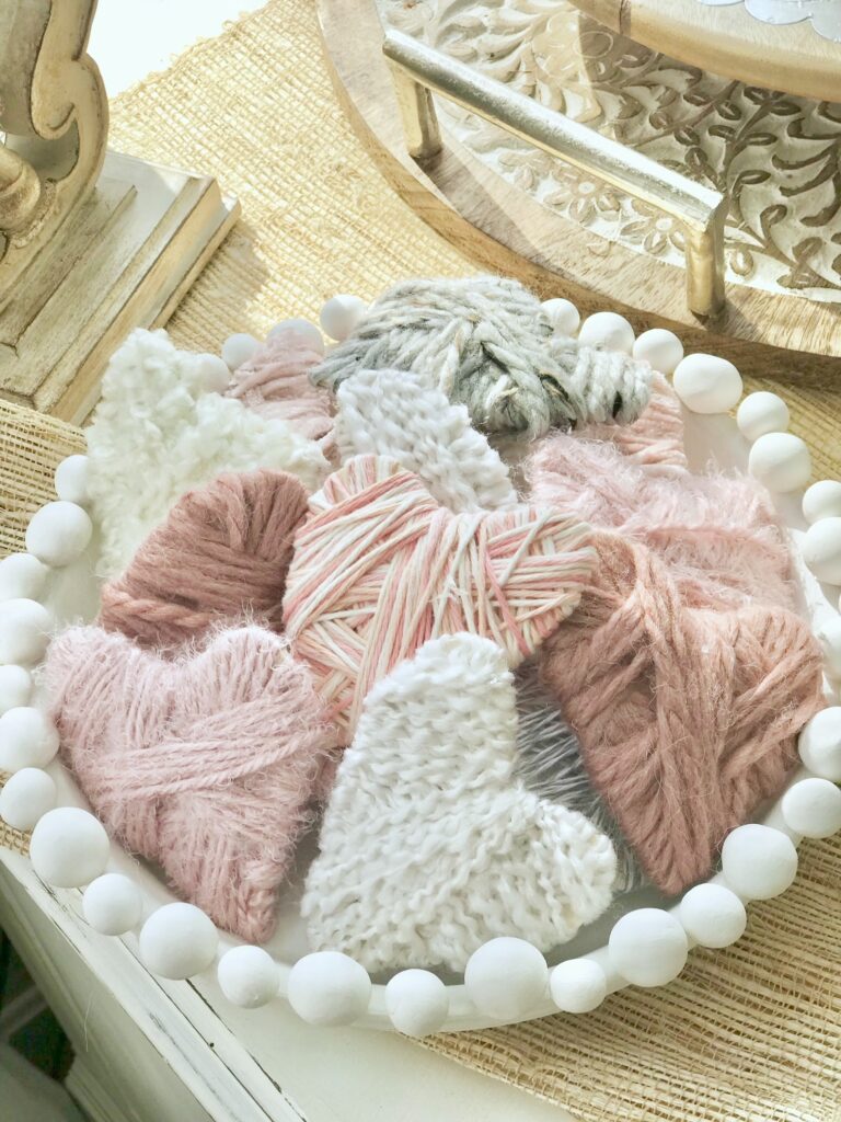 A white bowl with a collection of yarn wrapped hearts in pink, white, mauve and gray. 