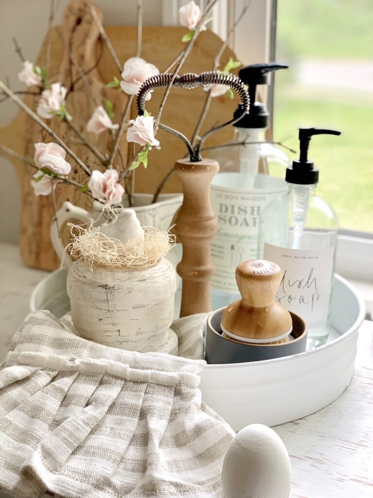 A kitchen sink tray that is white with scrub brushes, dish soap and bird and cherry blossom branches in a small pitcher. 