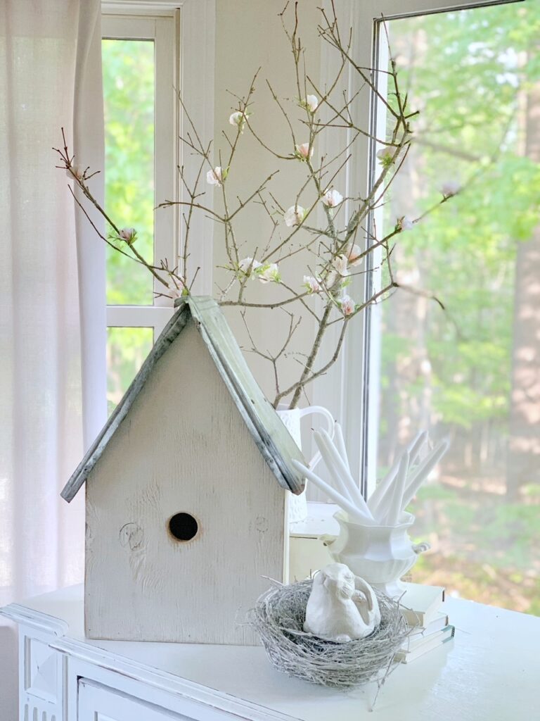 A large wooden white bird house in front of a white pitcher of faux cherry blossom branches.