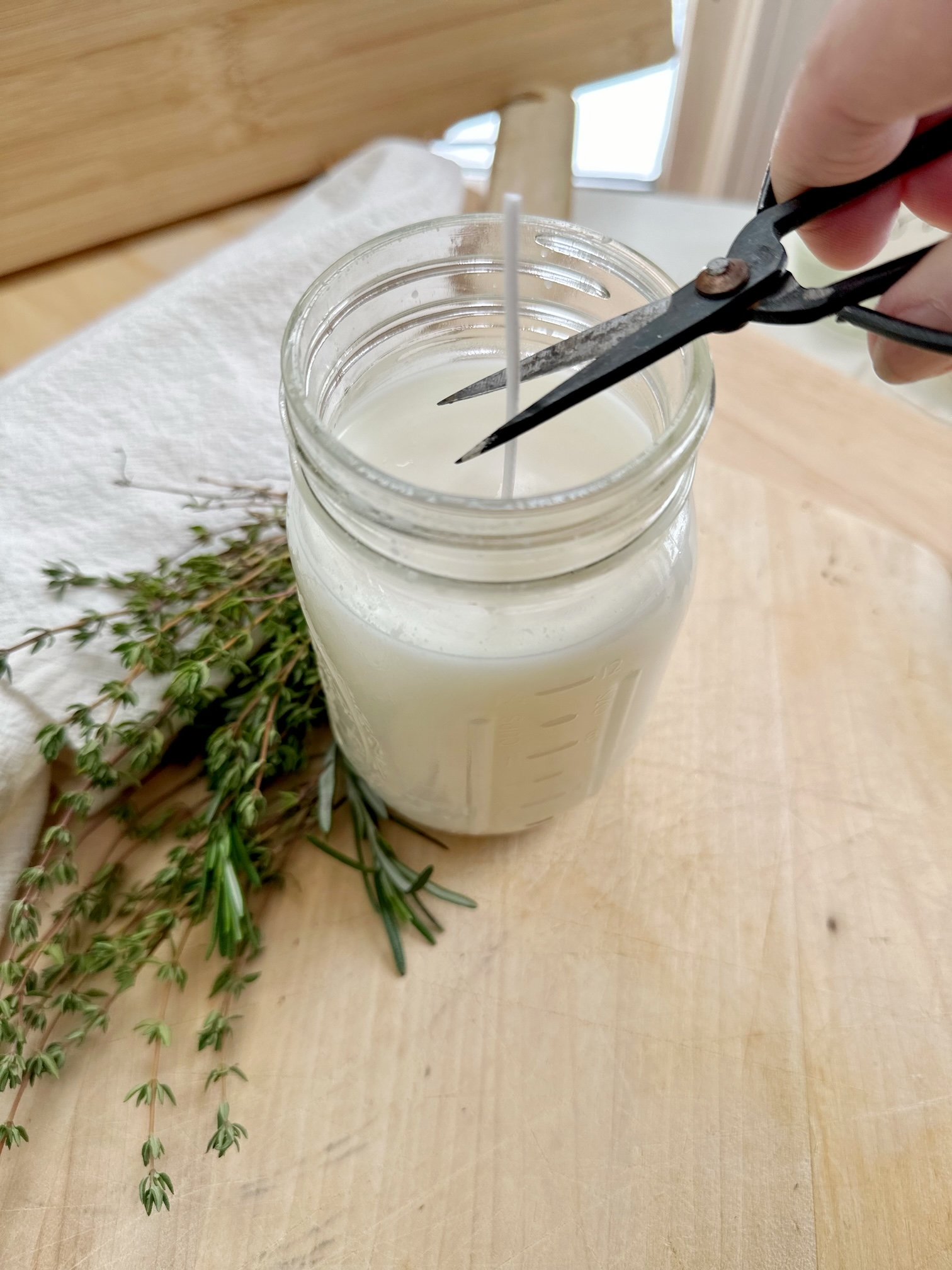 Cutting the wick of a candle. The candle is white and in a jar. There is a cluster of herbs next to it. 