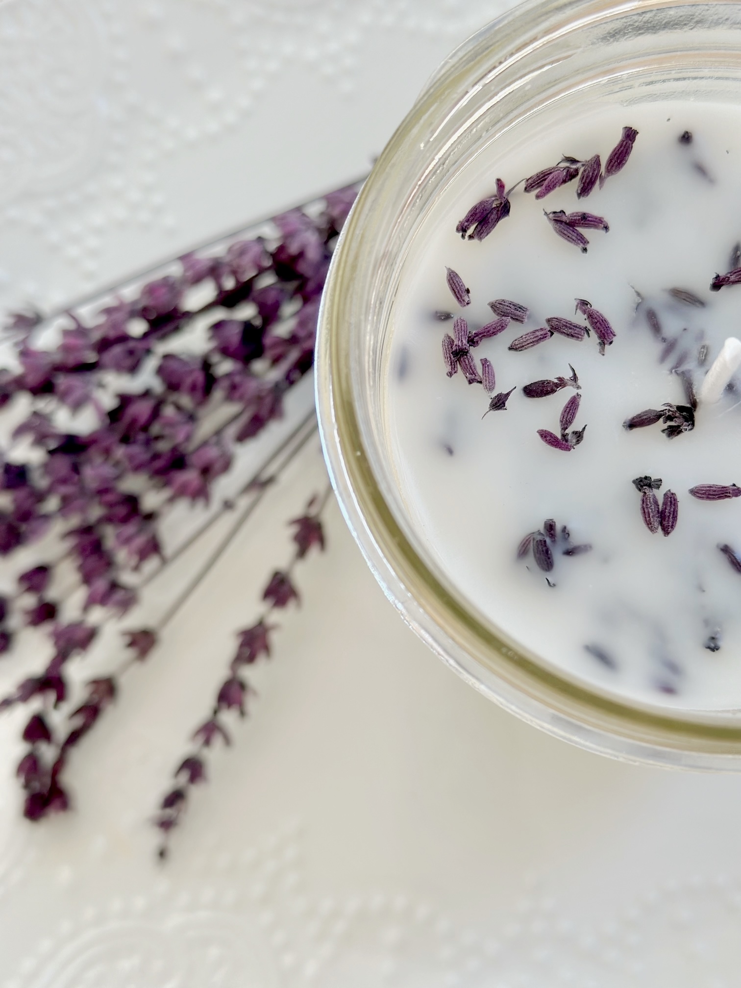 A top view of a lavender candle with a sprig of lavender laying next to it. the candle has lavender flowers (buds) sprinkled on top. 
