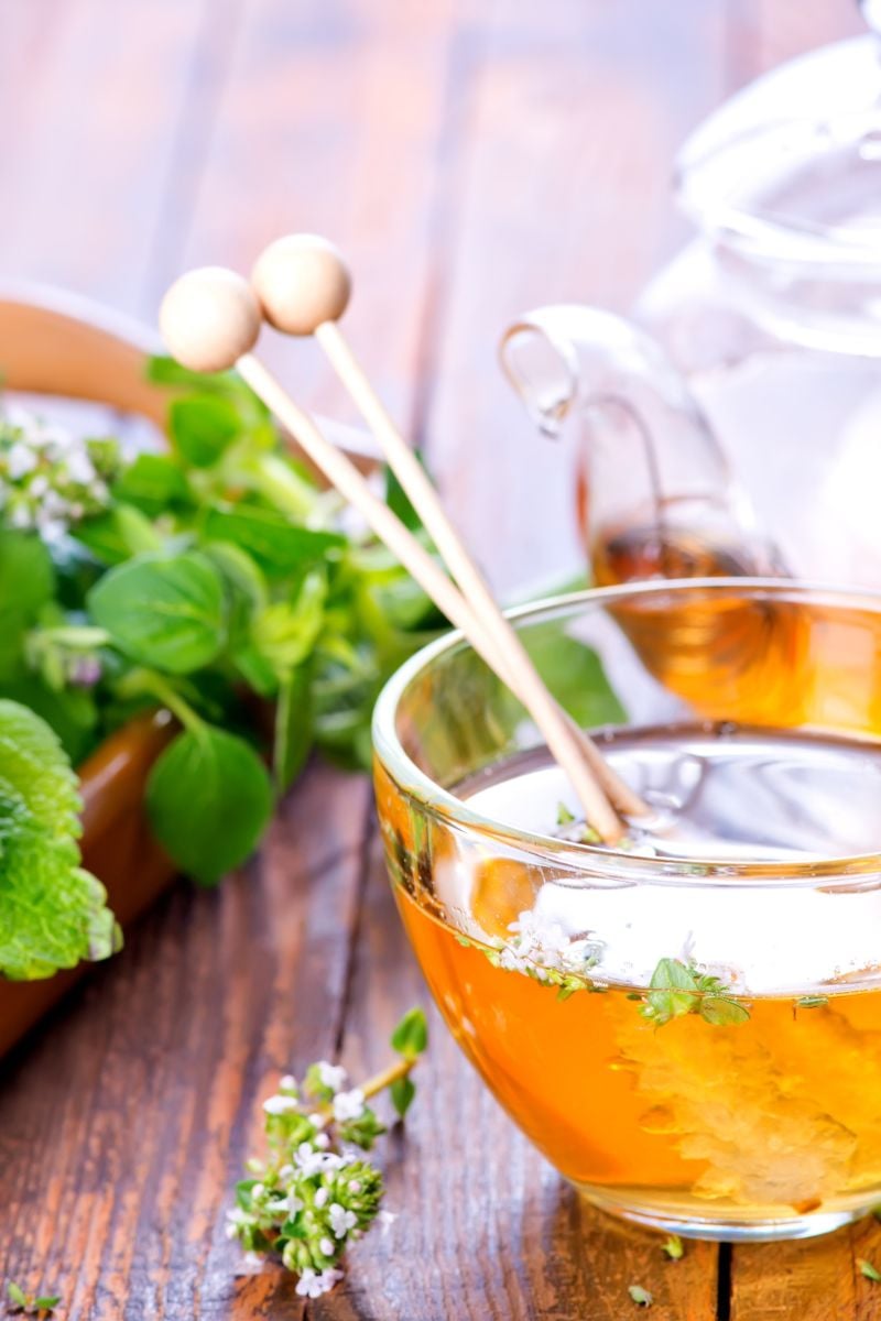 A clear tea cup with tea, stirrers and herbs. 