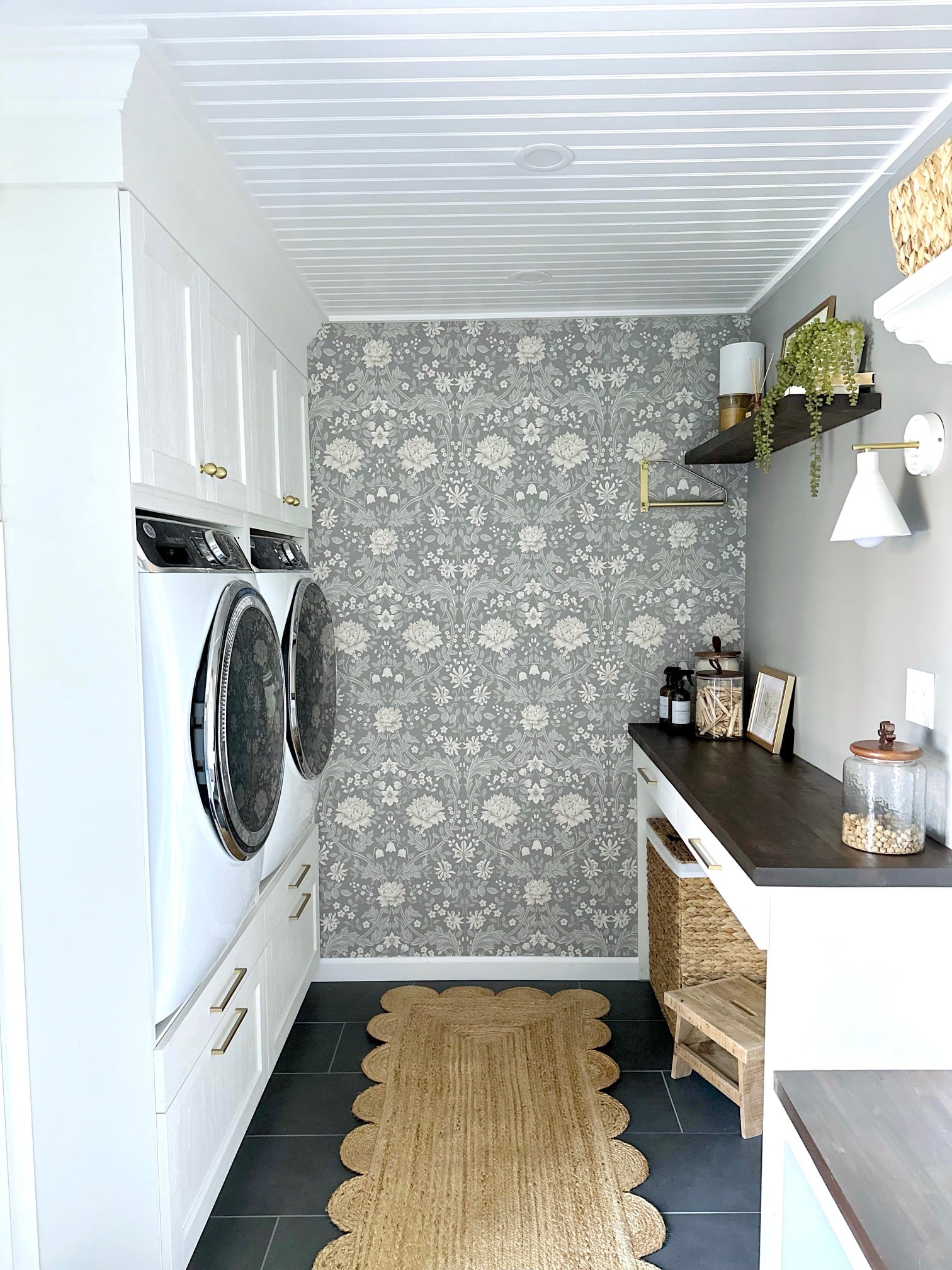 A view looking into the galley style laundry room with washer and dryer units raised in built in IKEA cabinets. on the right is a folding station as well as a dog walking station. the back wall has an accent wall with a lovely floral wallpaper with a gray background.
