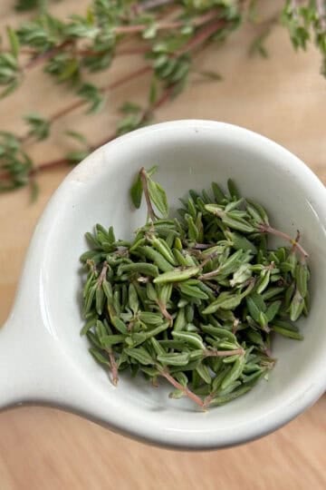 A tablespoon with thyme leaves in it. There are stems of fresh thyme in the background.