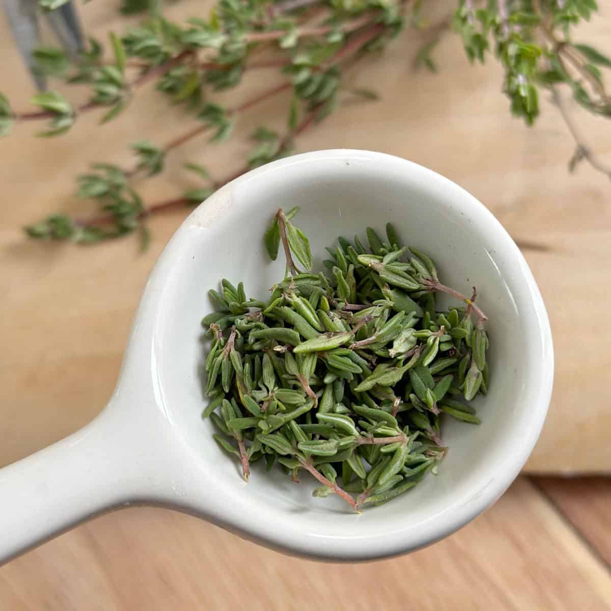 A tablespoon with thyme leaves in it. There are stems of fresh thyme in the background.