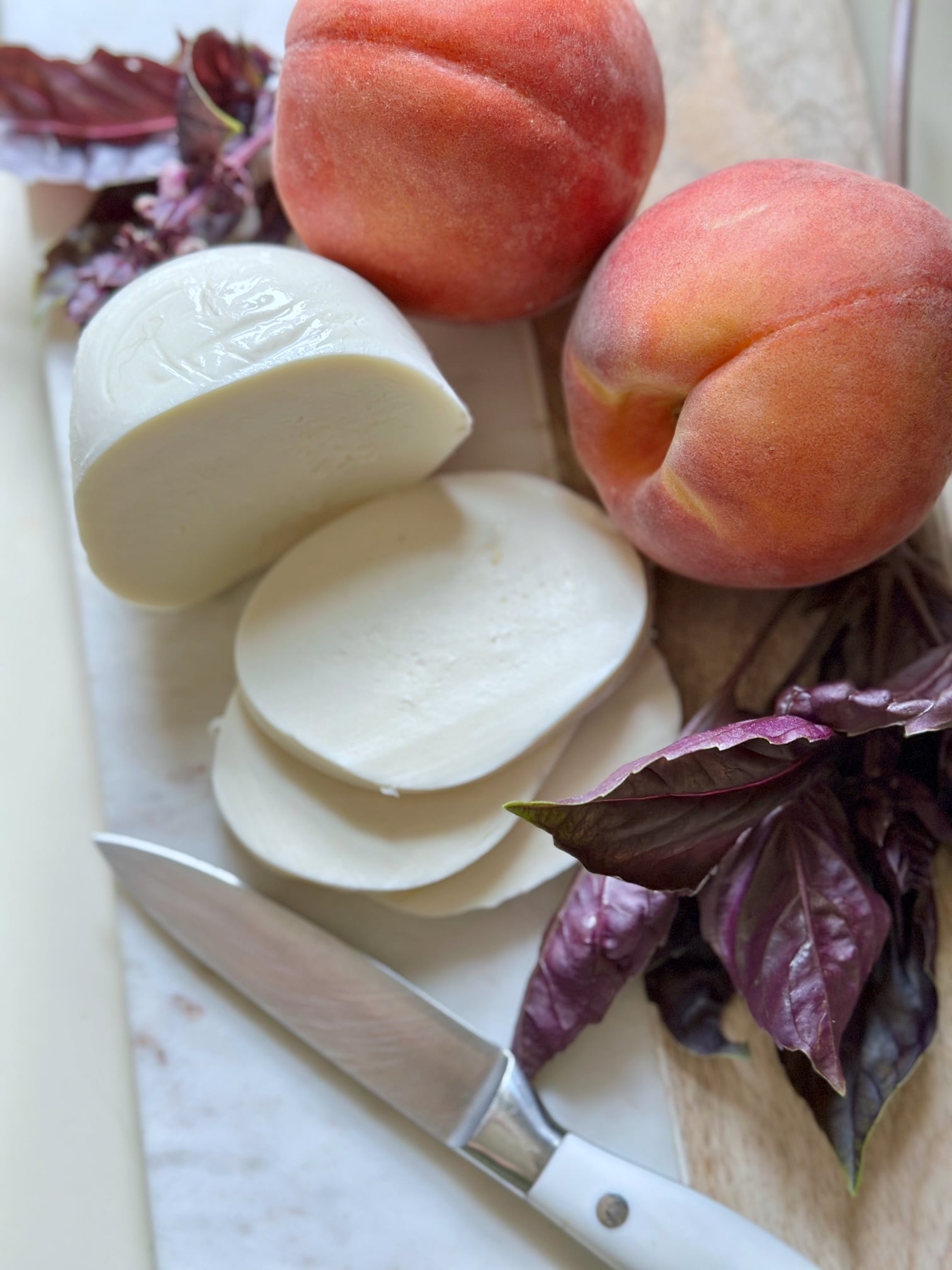 The ingredients, peaches, mozzarella cheese, purple basil on a cutting board with a pairing knife. 