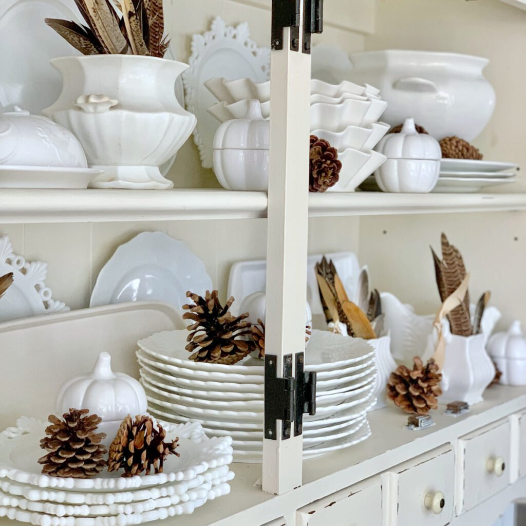 A fall themed hutch with all white plates, brown feathers, pears and pinecones.