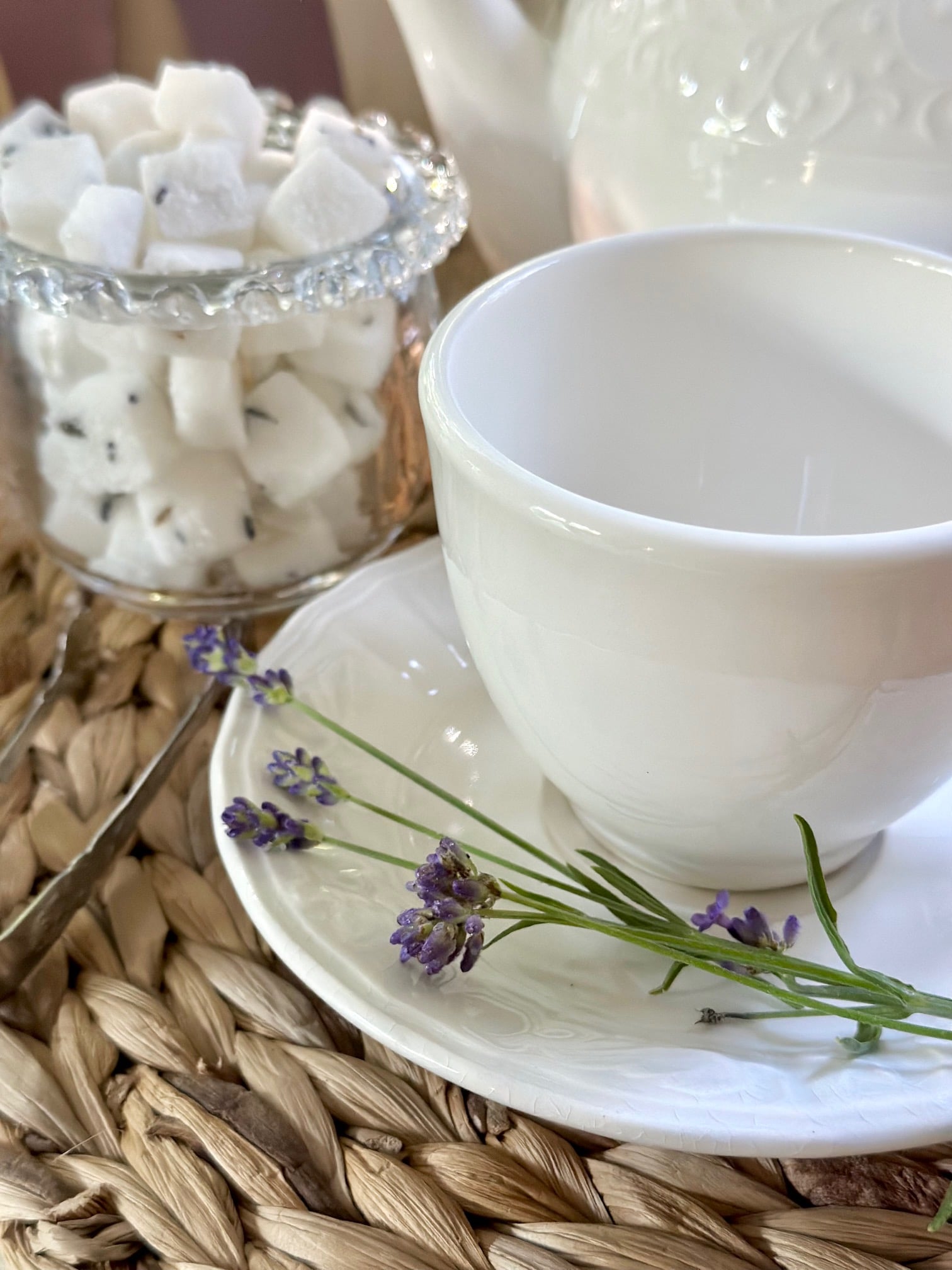 lavender sugar cubes in a clear glass sugar bowl behind a white tea cup with fresh lavender sprigs lying on the saucer. 