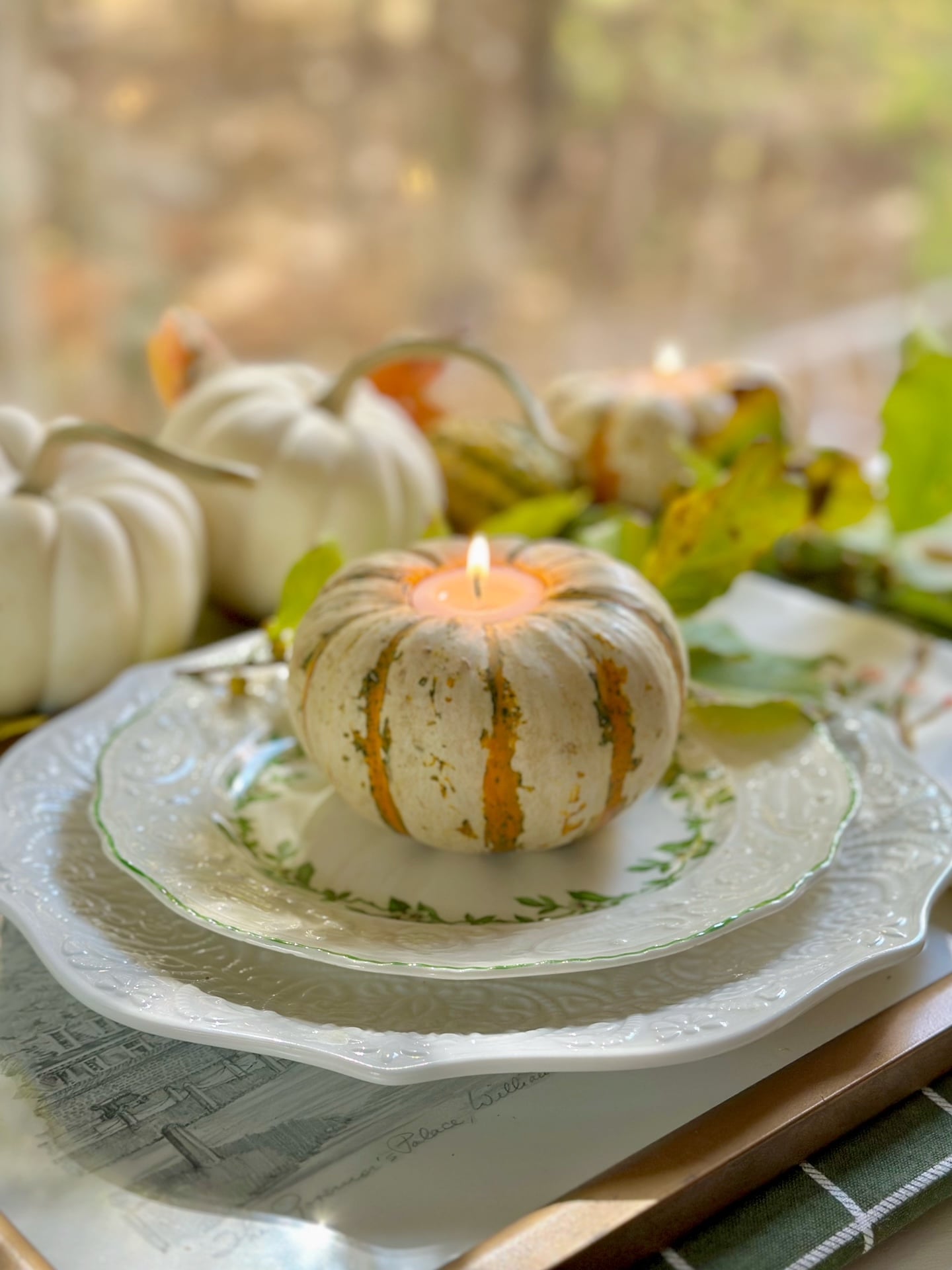 A tiger mini pumpkin that is mostly white with green and orange stripes with a white votive candle sunken into the center. This candle in a pumpkin sits on a salad plate with fresh fall branches and other mini pumpkins and gourds in the background.