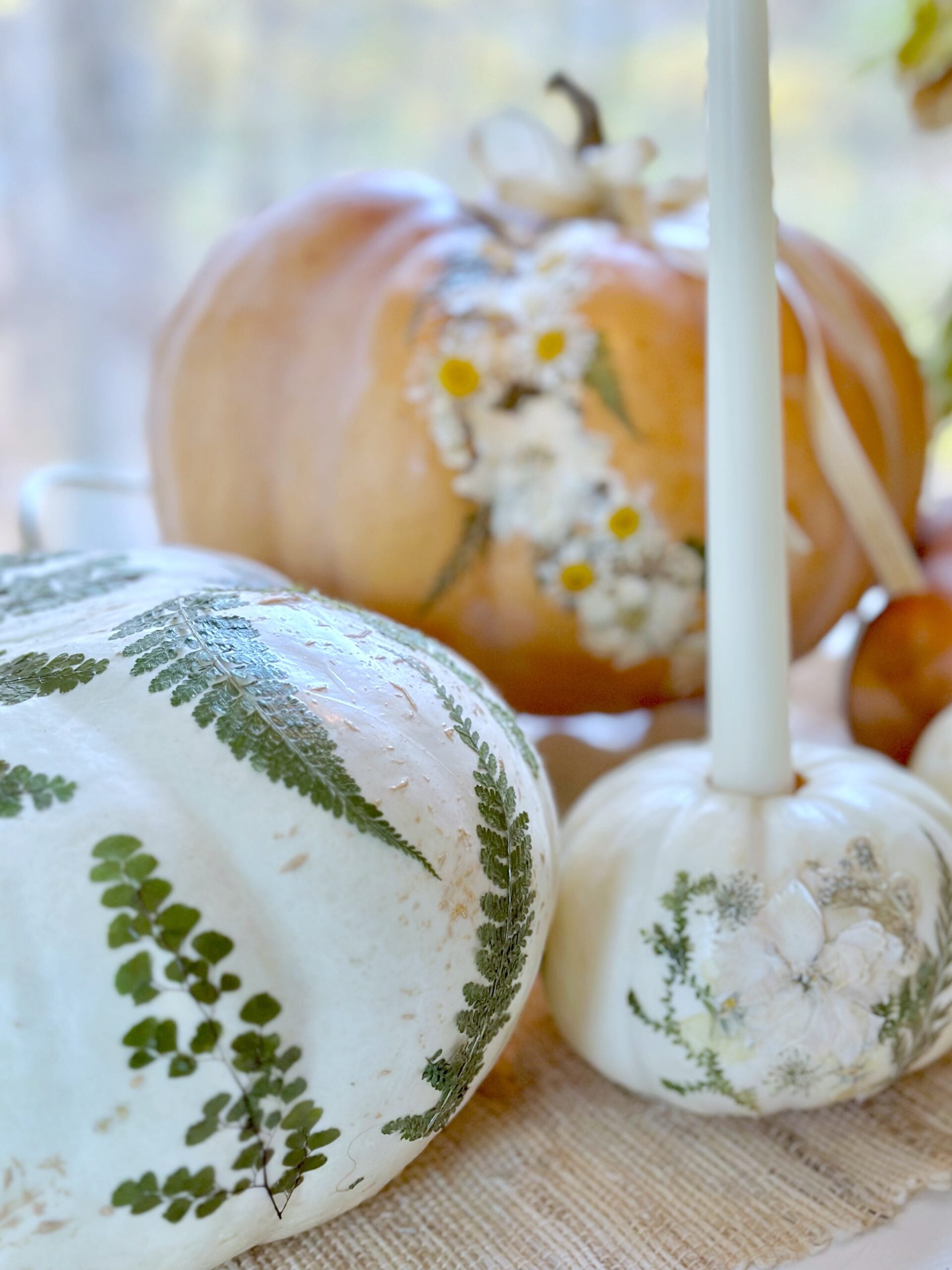 3 pressed flower pumpkins, 1 large white with just leaves. One small candle holder with white flowers and leaves and the last an orange pumpkin with flowers and greens cascading.