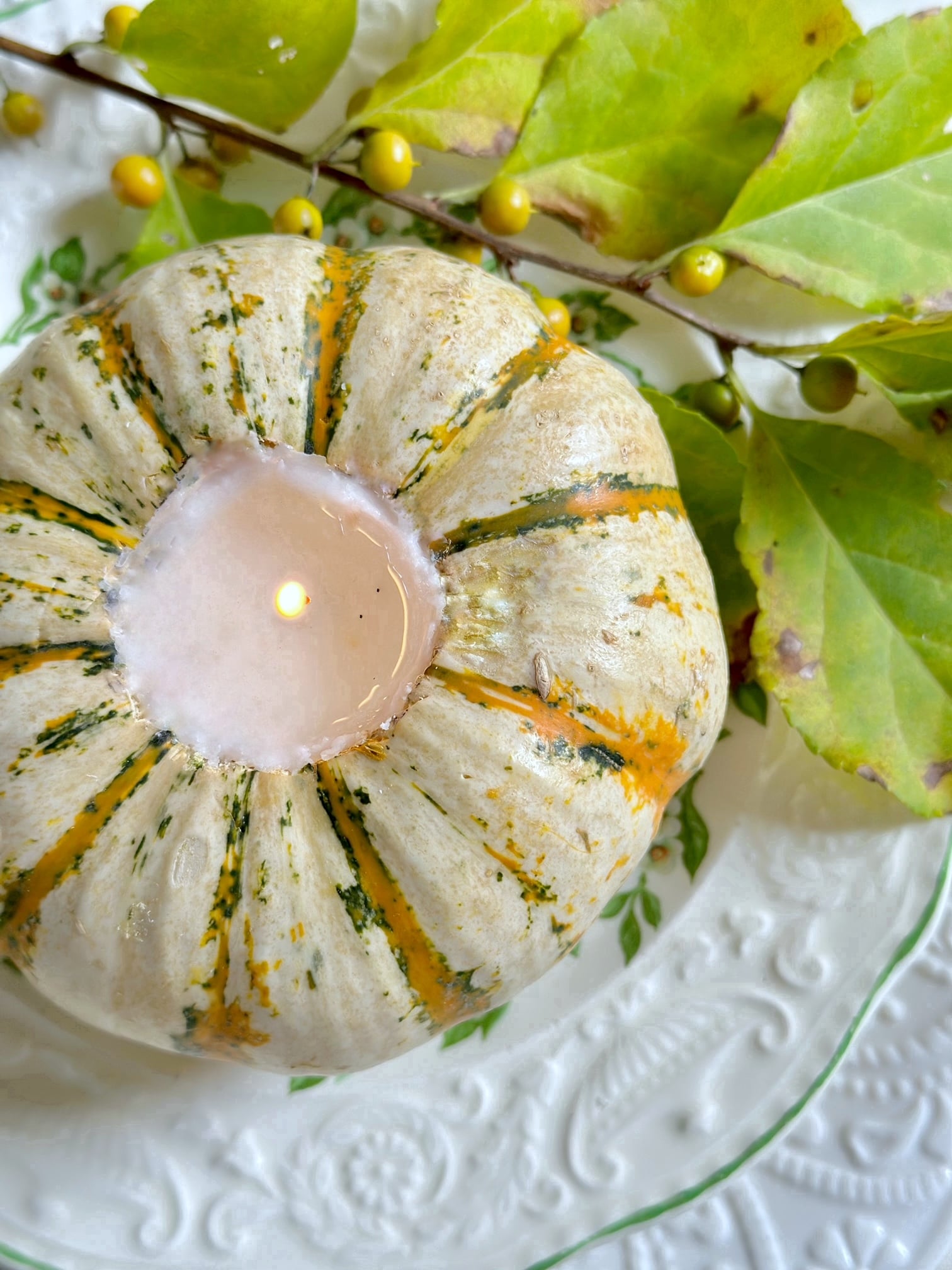 A top view of the pumpkin with the lit candle on a plate. there is a branch of yellow berries and green leaves as an accent next to the pumpkin.