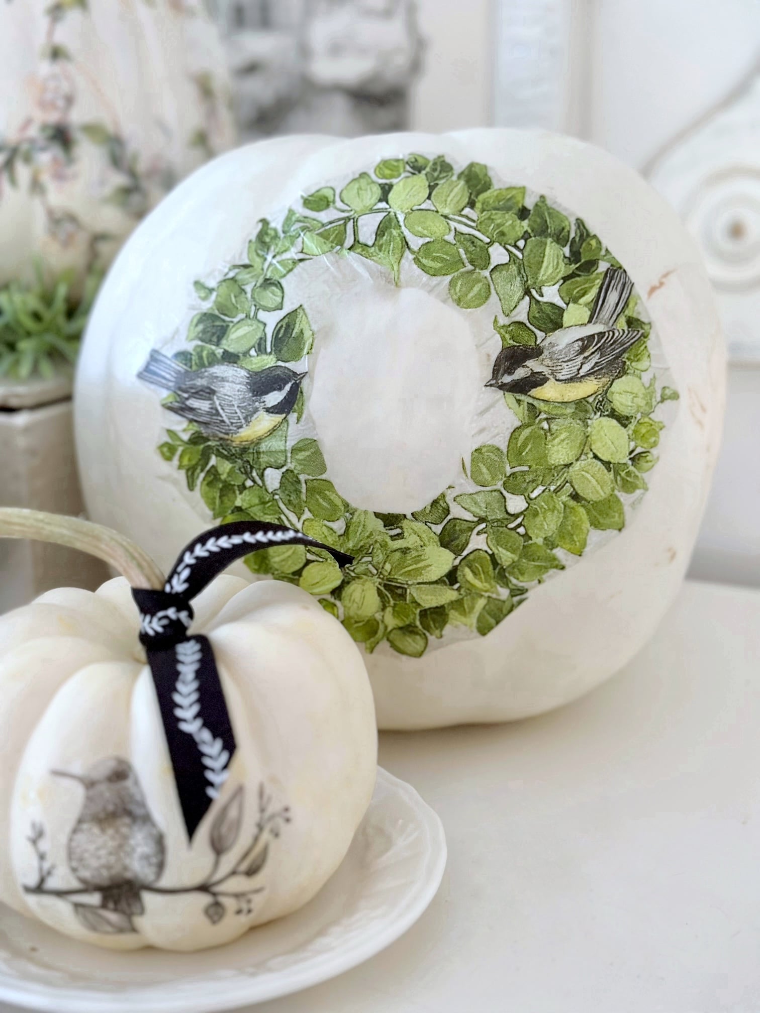 A large white pumpkin with a wreath transfer on the front. the wreath is large with 2 birds. 