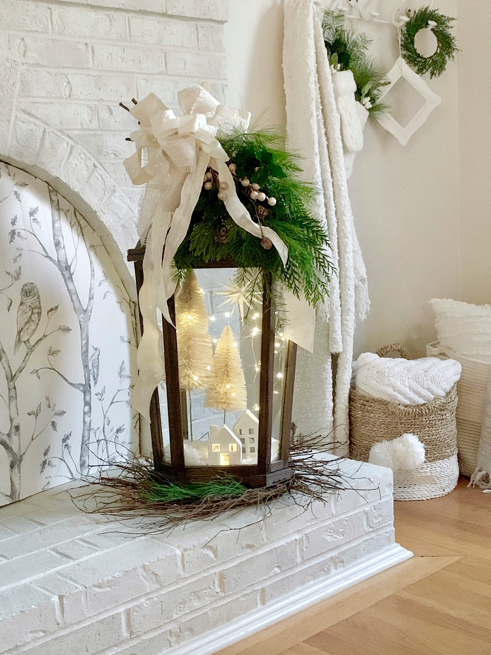 Large DIY Christmas Lantern on a white painted brick hearth. The lantern is wood and metal, has fresh green and large bow on top. Inside are twinkle lights, snow and a small village. 