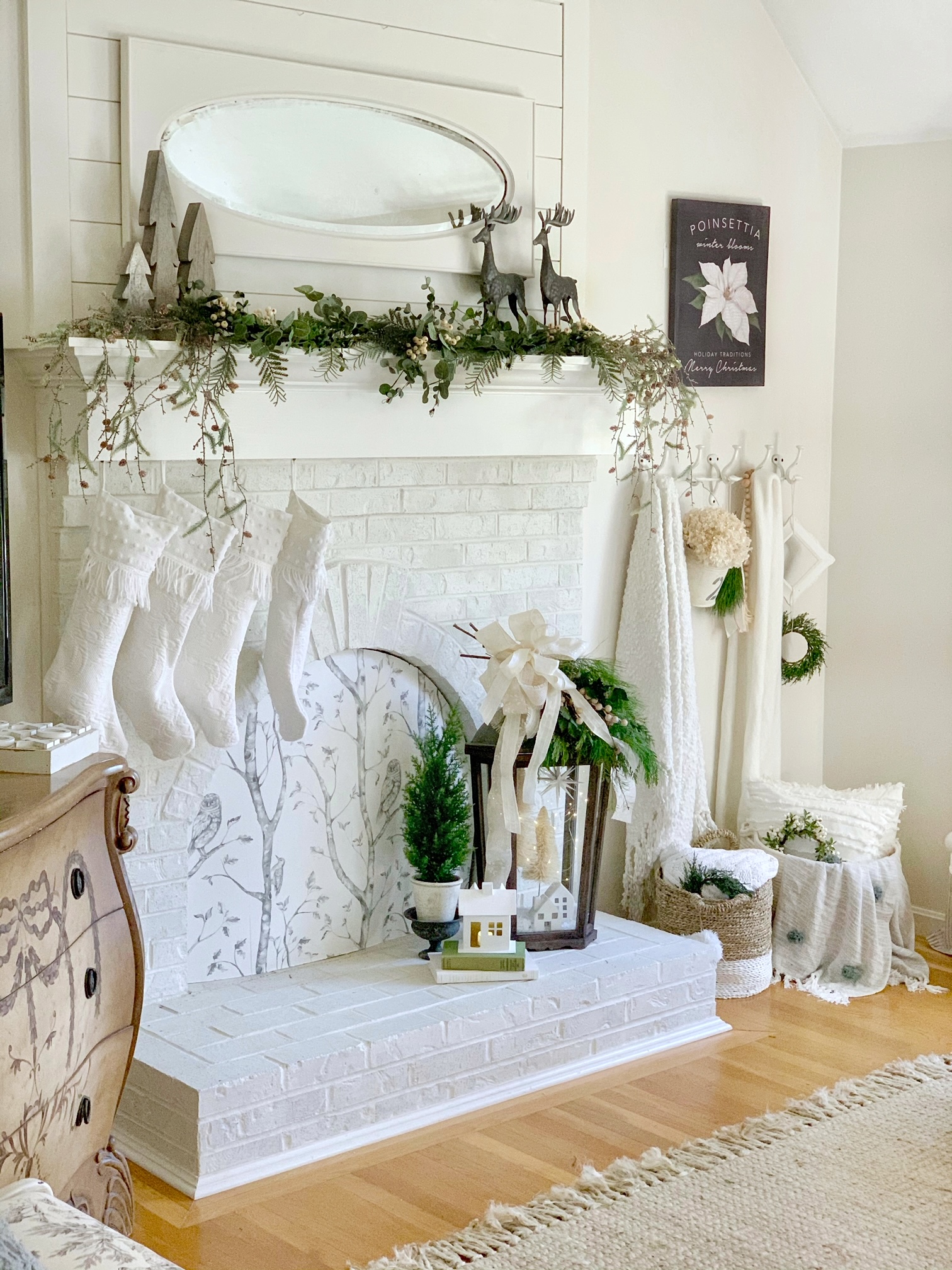 A white mantel with a fireplace cover board, stockings hanging, a decorated mantel. There are blankets hanging, baskets on the floor and the DIY Christmas Lantern is sitting on the hearth. 