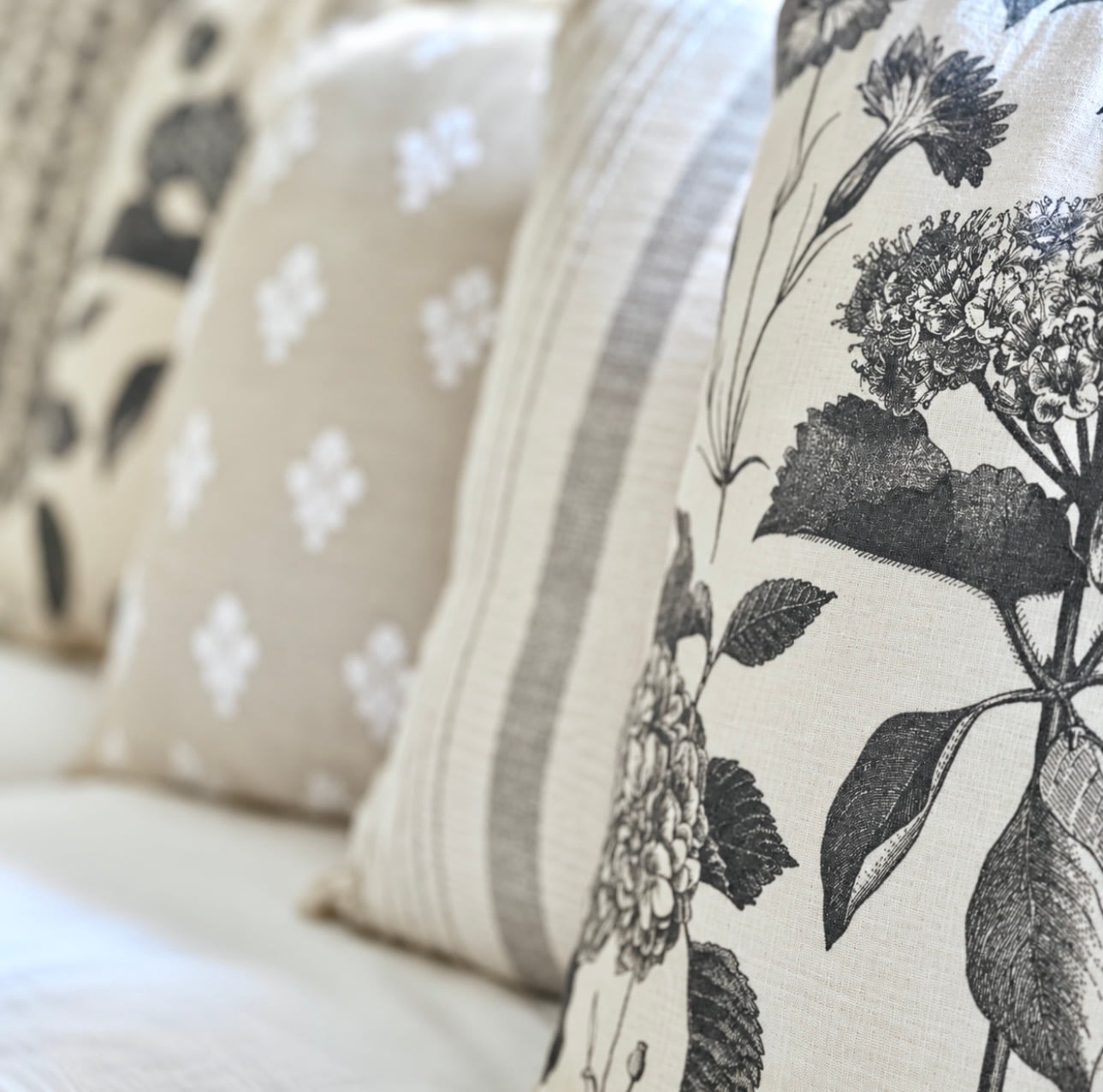 Four neutral throw pillows some florals, some stripes. Pillows are sitting on a white couch in a row.