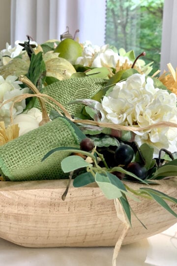 A dough bowl filled with fall items including pumpkins, ribbon, fruits , flowers and greens.