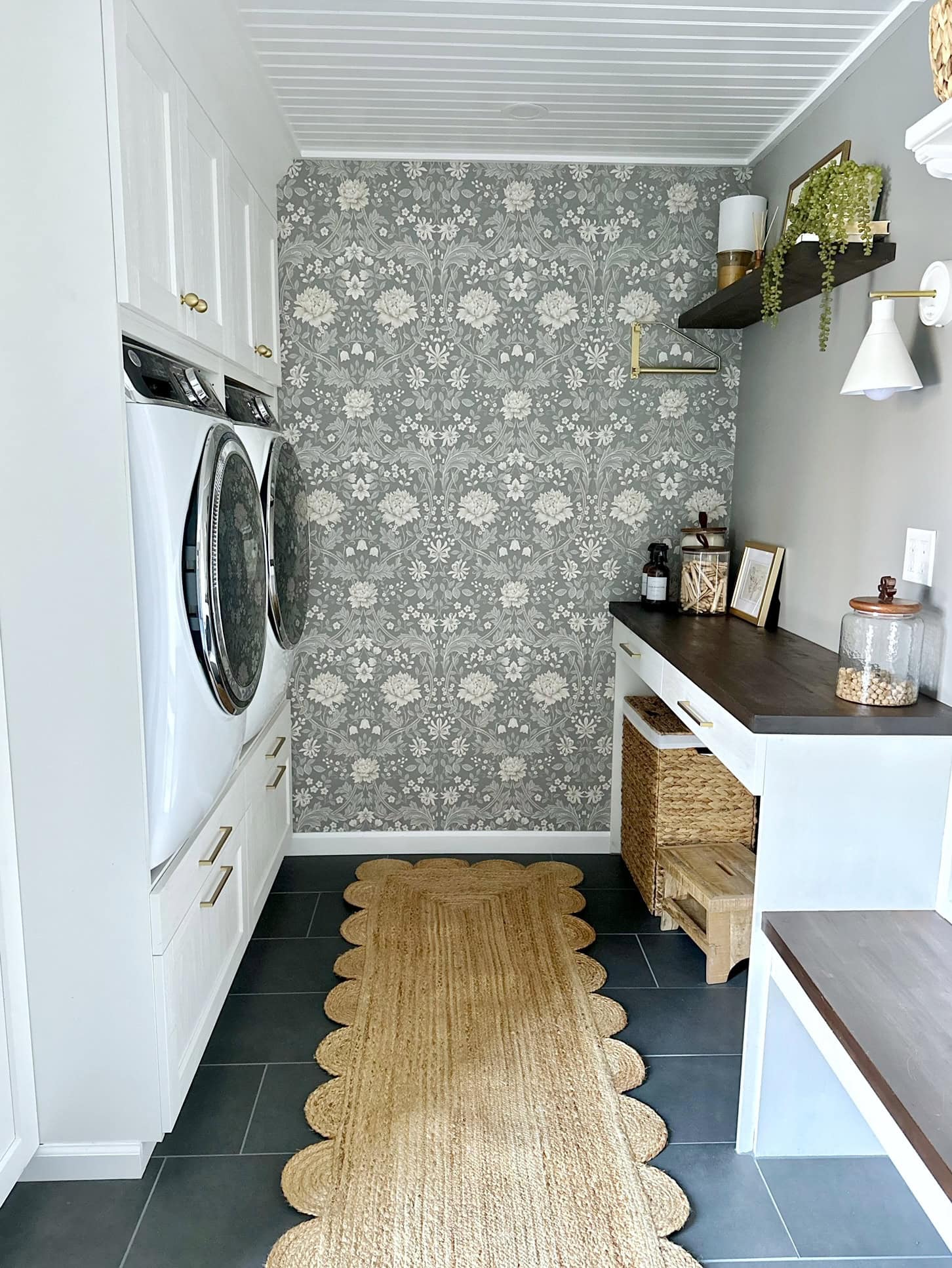 A full view of our laundry room. Its set up much like a galley kitchen. At the end of the room is a wall with wallpaper. the wallpaper has a gray background and is floral. 