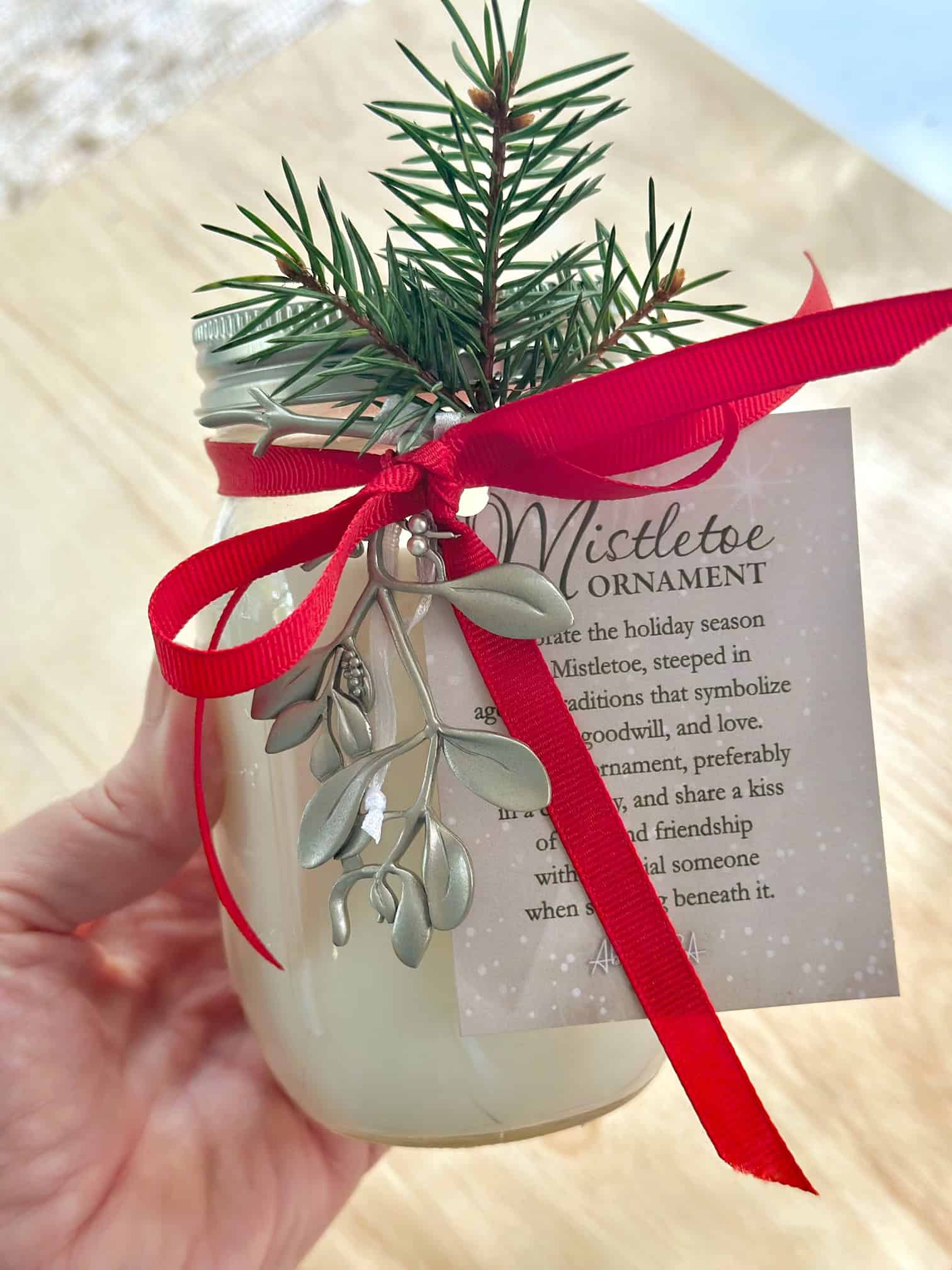 A beautiful candle made in a jar. The candle is mistletoe scented and has a red bow with some greenery. There is a silver mistletoe ornament hanging off the jar.