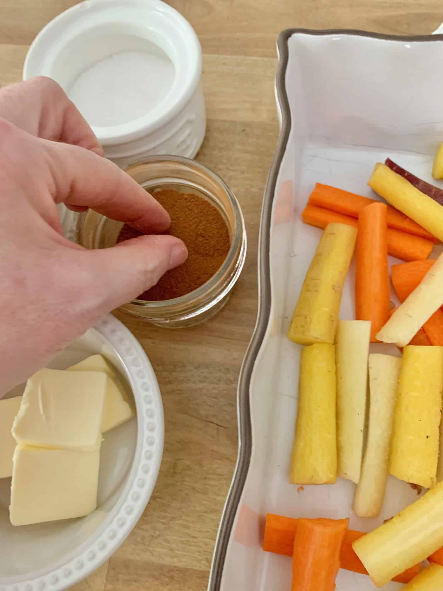 Pinching cinnamon from a spice jar to sprinkle on cut carrots in a baking dish.