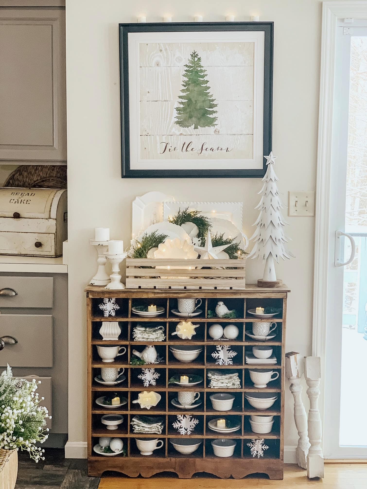 A vintage cubby filled with small cups, bowls, candle, and paper snowflakes.