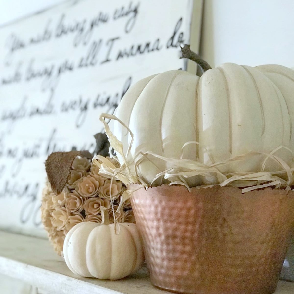 A large white faux pumpkin, sitting in a copper hammered planter with raffia under it. 
